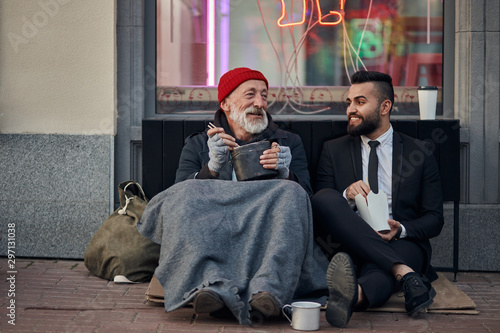 Handsome businessman in suit sitting on floor with homeless man together, listen to his story of life. Contrast people, rich and poor, but doesn't matter photo