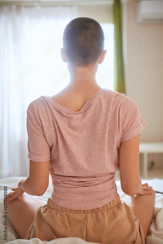 Back view of woman in t-shirt doing yoga exercise on bed in light room. Sit in pose lotus, meditating Back view of woman in t-shirt doing yoga exercise on bed in light room. Sit in pose lotus, meditating