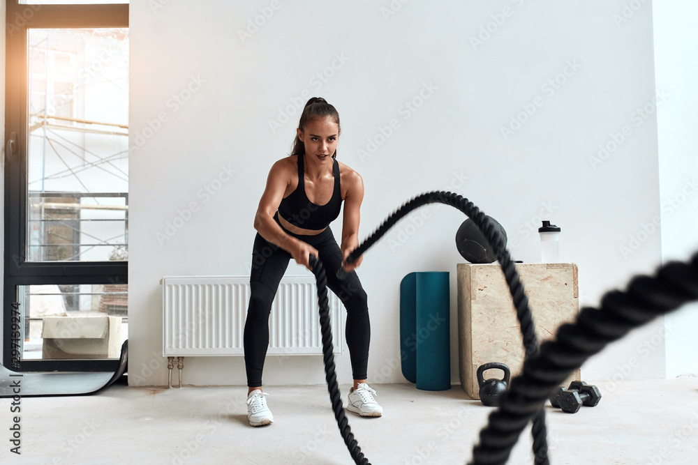Concentrated Caucasian female doing crossfit exercises using rope in light gym. Young beautiful woman in sportswear