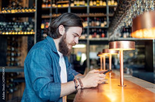 Young brown-haired bearded man using smartphone by a bar