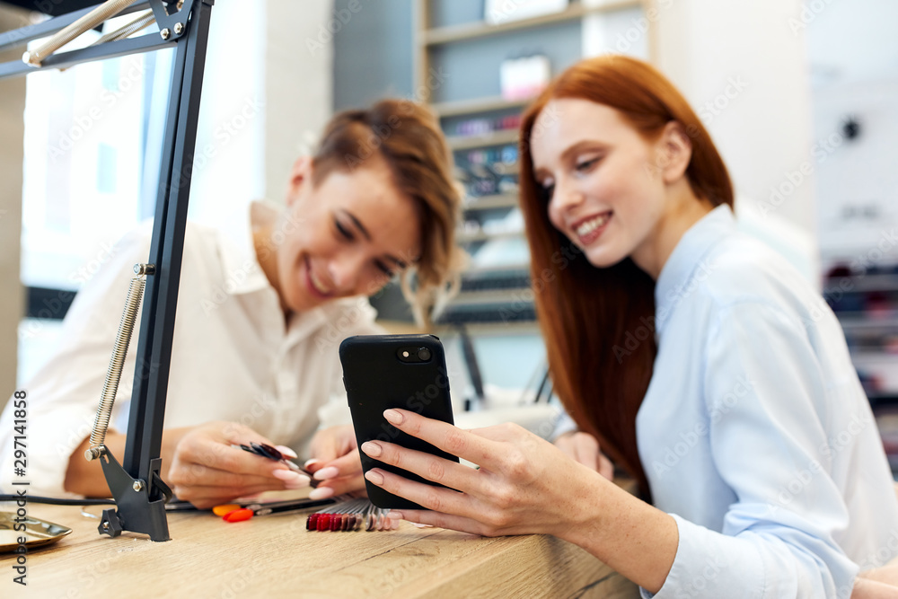 Smiling master and client look at phone on beautiful manicure. Redhead girl want manicur like on smartphone.