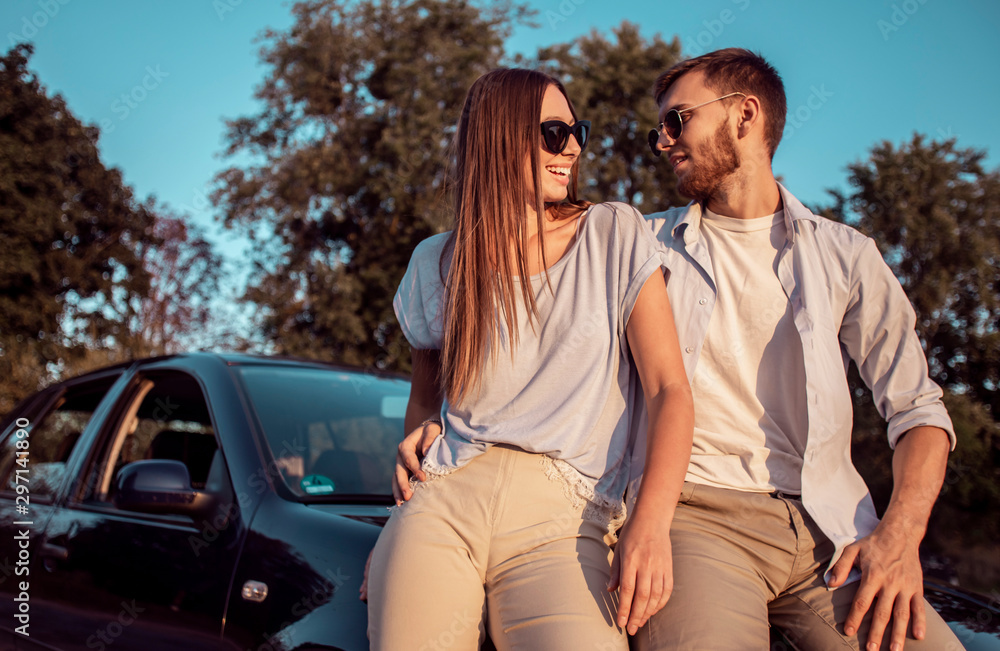Young man and woman talking while sitting on a car hood on a countryside road