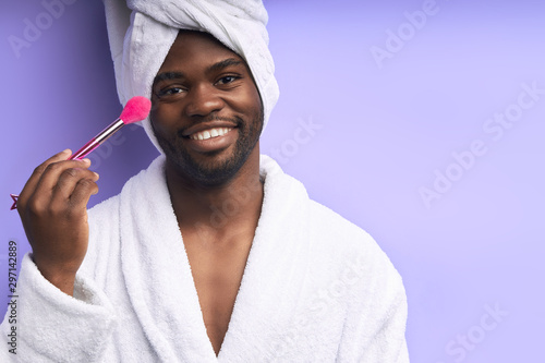 Positive smiling man in white bathrobe and towel look at camera, holding make-up brush isolated over purple background