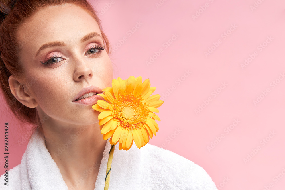 Young caucasian woman holding daisy yellow flower isolated over pink background. Female wearing white bathrobe, curlers on hair after shower. Natural beauty
