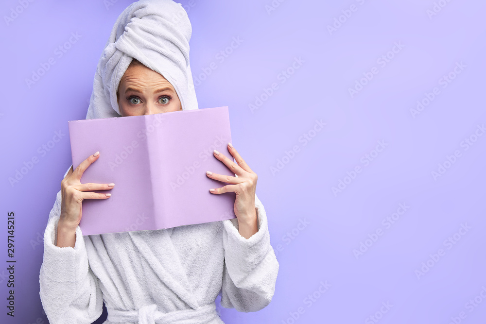 Open-eyed woman wearing bathrobe and towel after shower reading book isolated over purple background. Emotions, reading Open-eyed woman wearing bathrobe and towel after shower reading book isolated over purple background. Emotions, reading