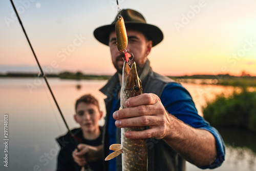 Positive man with teenager boy standing together and showing catch fish outdoors