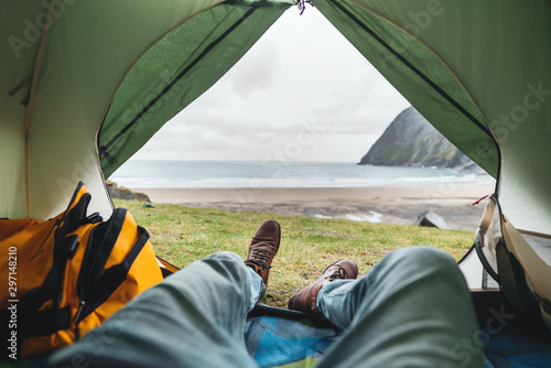 POV view from tent on on legs wearing hiking shoes in front of mountains and ocean. Alone tourist resting in camping by the sea