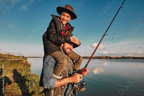 Son and father have fun while fishing. Father holds son on shoulders. Boy hold head. They smile. Background lake.