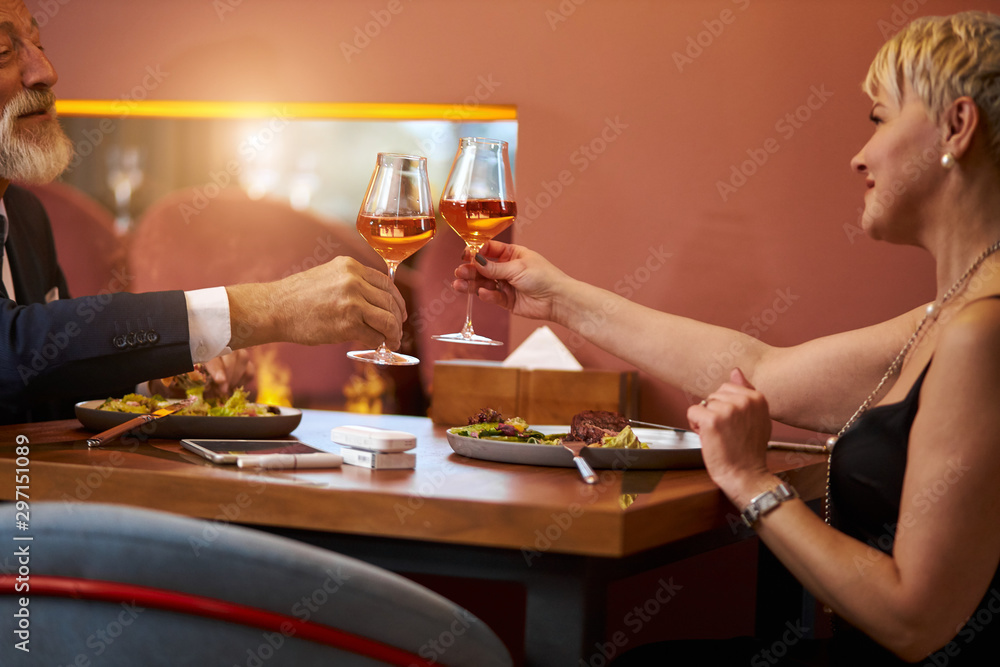 Grey-haired man in tuxedo and senior lady in black dress clink glasses with champagne in expensive restaurant. Look and happily smile. Romantic atmosphere