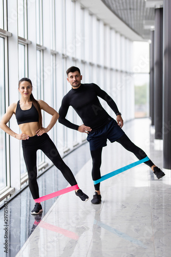Fototapeta Naklejka Na Ścianę i Meble -  Young Couple standing and keeping resistance band on hips wearing sportswear and trainers at gym