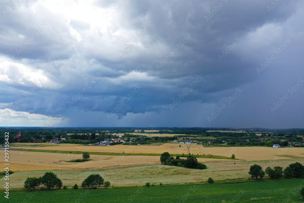 Aerial drone perspective view on dark storm clouds over the green scenery with green forests, yellow wheat fields, green meadows and villages