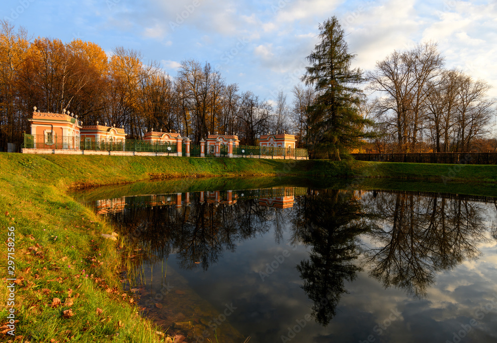 Beautiful view of Kuskovo park in Moscow at sunny autumn evening with lush grass, yellow leaves and pond with reflection of blue cloudy sky, pine tree and bird houses. Kuskovo, Moscow Beautiful view of Kuskovo park in Moscow at sunny autumn evening with lush grass, yellow leaves and pond with reflection of blue cloudy sky, pine tree and bird houses. Kuskovo, Moscow