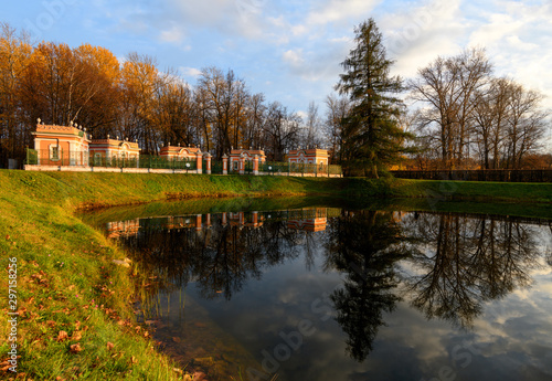 Beautiful view of Kuskovo park in Moscow at sunny autumn evening with lush grass, yellow leaves and pond with reflection of blue cloudy sky, pine tree and bird houses. Kuskovo, Moscow