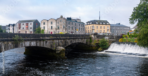 Beautiful view of the River Corrib flowing through the center of Galway city, with old buildings and wild flowers growing on a sunny summer day.