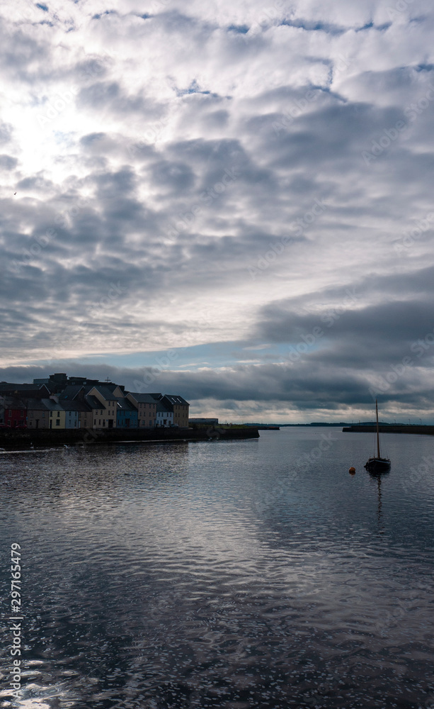 One sail boat on a calm Galway bay with the famous colorful houses & sunrise, ocean & cloudy sky in the distance. Taken early morning.