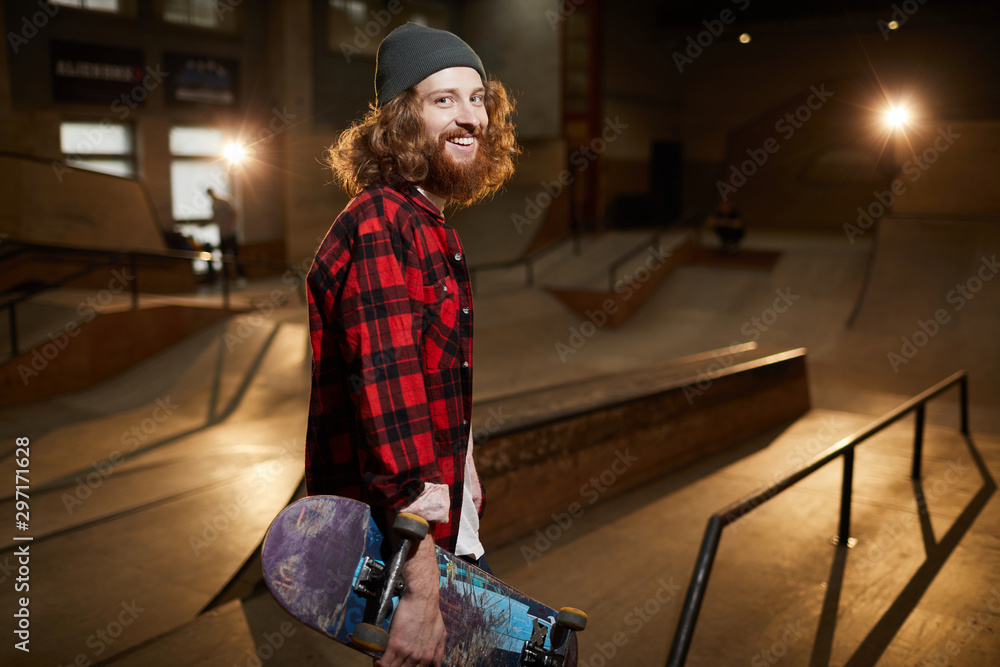 Waist up portrait of contemporary bearded skater looking at camera and smiling while posing in urban skating park lit by dim light, copy space