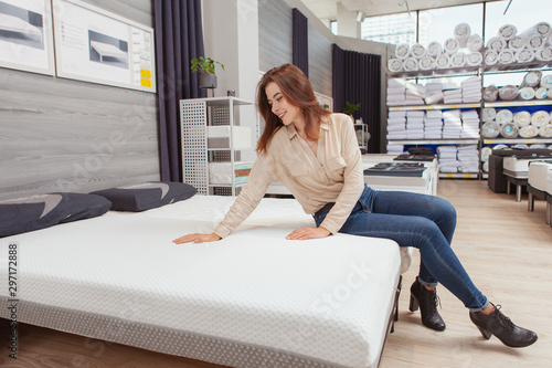Attractive female customer sitting on a new bed at furniture store, choosing orthopedic mattress to buy photo