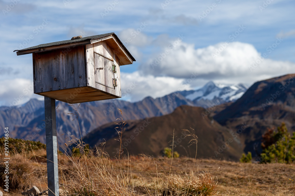 A small handmade wooden bird house on a pole standing on top of a mountain in the alps. In the background summits covered in snow are viisible.
