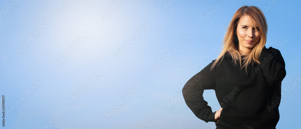 Portrait of a smiling beautiful girl in black hoodie standing isolated on light blue banner background Portrait of a smiling beautiful girl in black hoodie standing isolated on light blue banner background