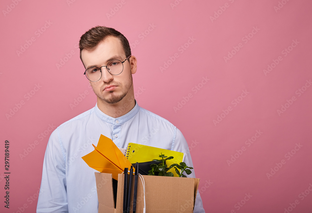 A pretty guy in a sky blue shirt and computer glasses stands on a pink background with a cardboard box with different things in his hands. Dismissal. Fired. A pretty guy in a sky blue shirt and computer glasses stands on a pink background with a cardboard box with different things in his hands. Dismissal. Fired.