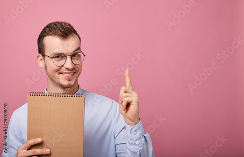 Fotomural A young student in a gently blue shirt with glasses holds in his hand a brown loose-leaf notebook and looks into the frame with smile, is on a pink background