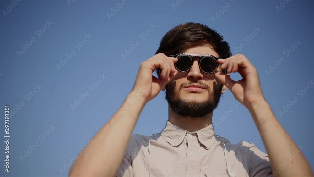 Portrait of serious bearded hipster man, wearing round sunglasses and shirt, on background of blue sky. Portrait of serious bearded hipster man, wearing round sunglasses and shirt, on background of blue sky.