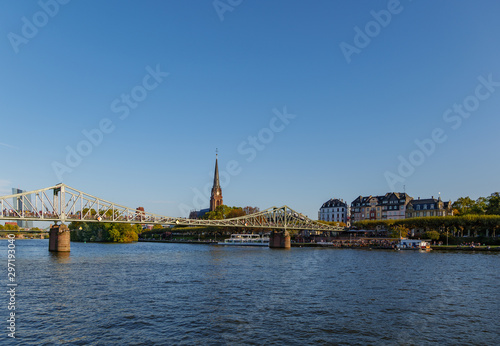 Outdoor sunny view of Eiserner Steg, historical pedestrian Iron Bridge, and promenade on riverside of Main River in sunny day in Frankfurt, Germany. © Peeradontax