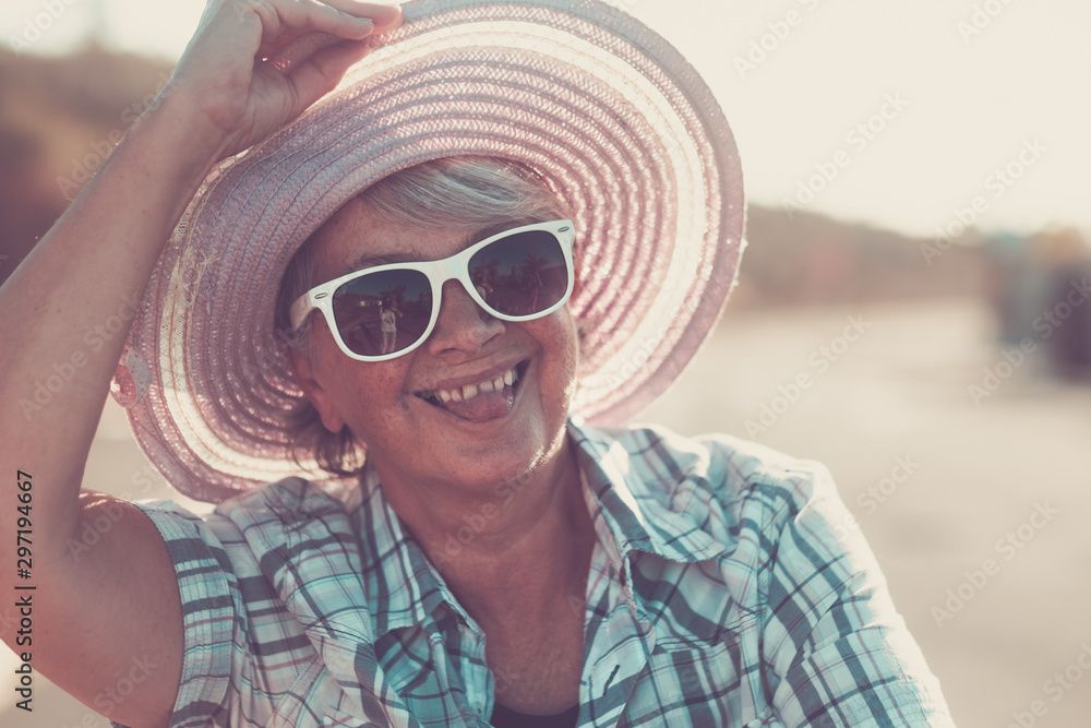 A senior woman stops riding her bicycle to look at the camera while making a tongue. A happy person with a big hat who enjoys nature and a healthy lifestyle. A senior woman stops riding her bicycle to look at the camera while making a tongue. A happy person with a big hat who enjoys nature and a healthy lifestyle.