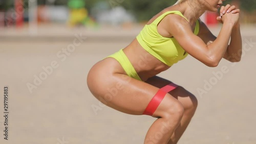 Resistance band fitness woman doing squat exercise for glutes on the beach. Athletic girl workout with fitness loop band outdoors