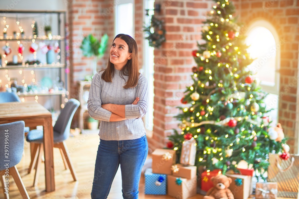 Young beautiful woman standing with happy face around christmas tree and presents smiling for festive celebration Young beautiful woman standing with happy face around christmas tree and presents smiling for festive celebration