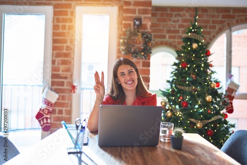 Beautiful woman sitting at the table working with laptop at home around christmas tree showing and pointing up with fingers number three while smiling confident and happy.