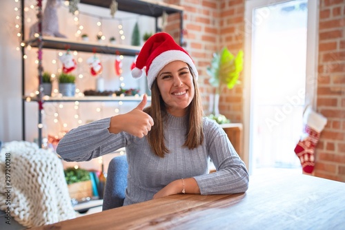 Young beautiful woman wearing christmas hat sitting at the table at home doing happy thumbs up gesture with hand. Approving expression looking at the camera with showing success.