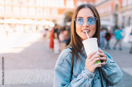 Happy urban woman with blue sunglasses enjoying her morning drinking a soda in styrofoam cup with straw. Pretty girl in the street. Take away beverages concept.