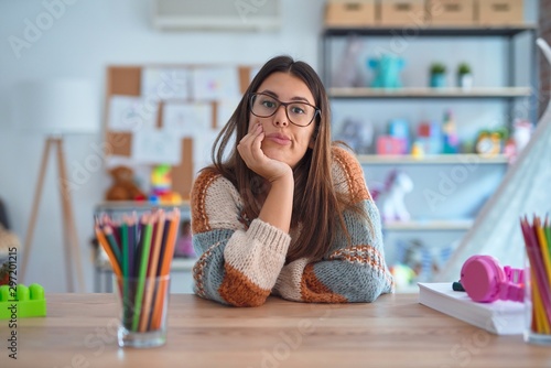 Quadro su tela Young beautiful teacher woman wearing sweater and glasses sitting on desk at kindergarten thinking looking tired and bored with depression problems with crossed arms