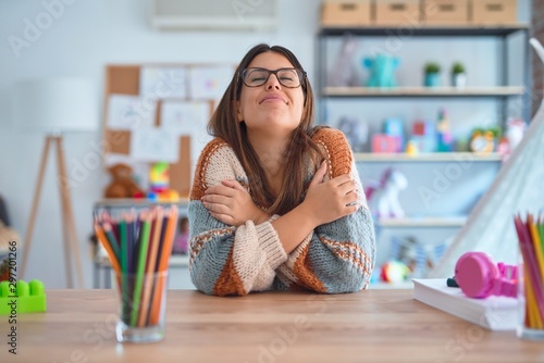 Young beautiful teacher woman wearing sweater and glasses sitting on desk at kindergarten Hugging oneself happy and positive, smiling confident. Self love and self care