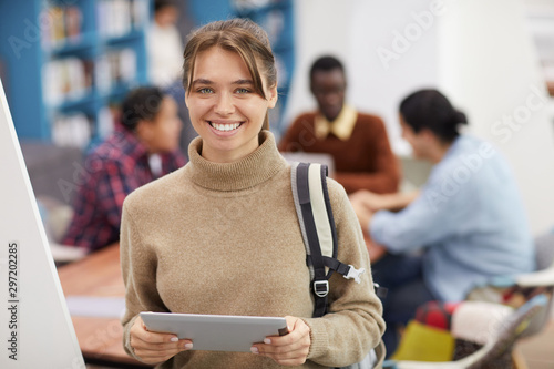 Waist up portrait of beautiful young woman smiling at camera while standing in library of modern college, copy space