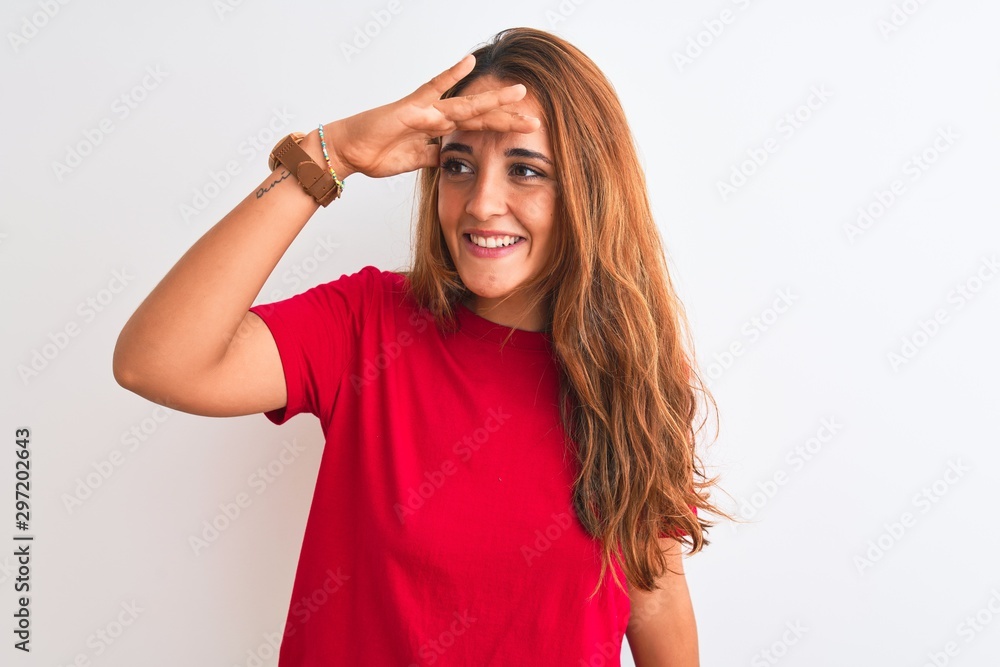 Young redhead woman wearing red casual t-shirt stading over white isolated background very happy and smiling looking far away with hand over head. Searching concept. Young redhead woman wearing red casual t-shirt stading over white isolated background very happy and smiling looking far away with hand over head. Searching concept.