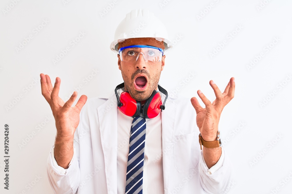 Young chemist man wearing security helmet and headphones over isolated background crazy and mad shouting and yelling with aggressive expression and arms raised. Frustration concept. Young chemist man wearing security helmet and headphones over isolated background crazy and mad shouting and yelling with aggressive expression and arms raised. Frustration concept.