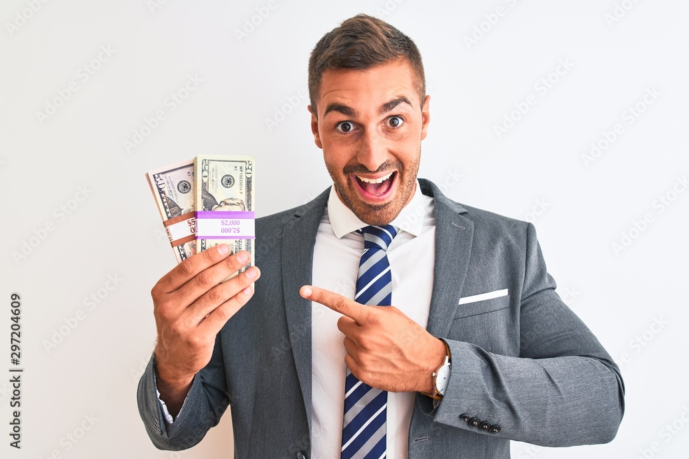 Young handsome business man holding bunch of dollars banknotes over isolated background very happy pointing with hand and finger Young handsome business man holding bunch of dollars banknotes over isolated background very happy pointing with hand and finger
