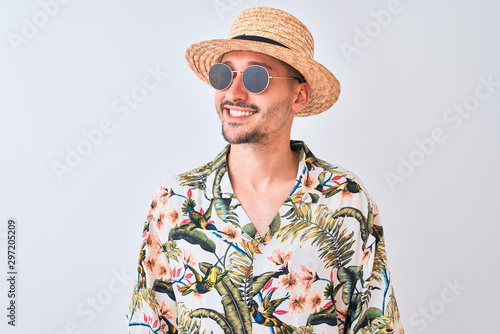 Young handsome man wearing Hawaiian shirt and summer hat over isolated background looking away to side with smile on face, natural expression. Laughing confident.