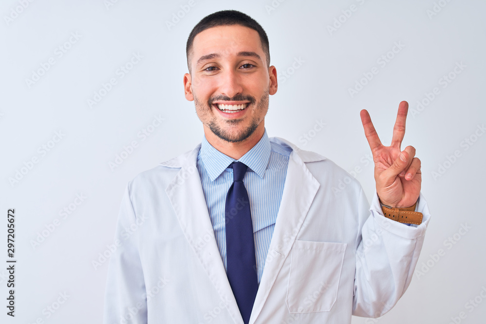 Young doctor man wearing white coat over isolated background smiling with happy face winking at the camera doing victory sign. Number two. Young doctor man wearing white coat over isolated background smiling with happy face winking at the camera doing victory sign. Number two.