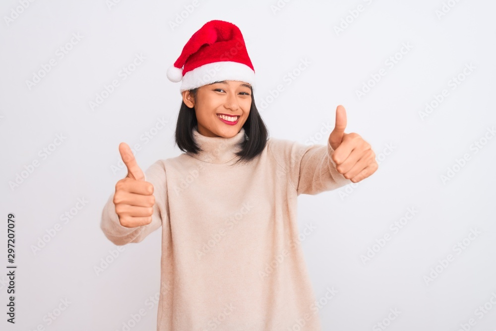 Young beautiful chinese woman wearing Christmas Santa hat over isolated white background approving doing positive gesture with hand, thumbs up smiling and happy for success. Winner gesture. Young beautiful chinese woman wearing Christmas Santa hat over isolated white background approving doing positive gesture with hand, thumbs up smiling and happy for success. Winner gesture.
