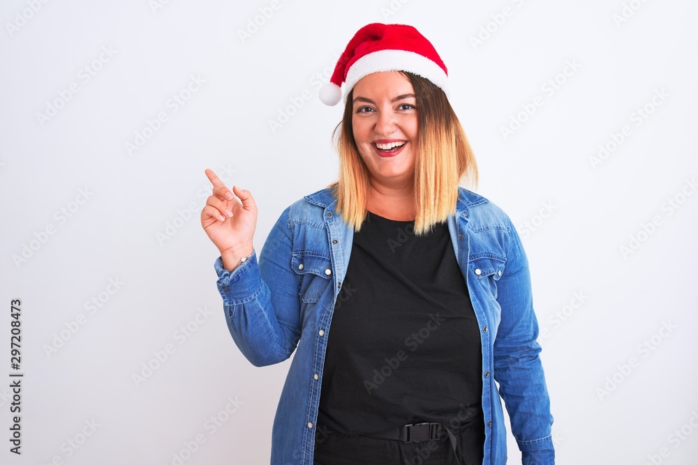 Young beautiful woman wearing Christmas Santa hat standing over isolated white background with a big smile on face, pointing with hand and finger to the side looking at the camera. Young beautiful woman wearing Christmas Santa hat standing over isolated white background with a big smile on face, pointing with hand and finger to the side looking at the camera.