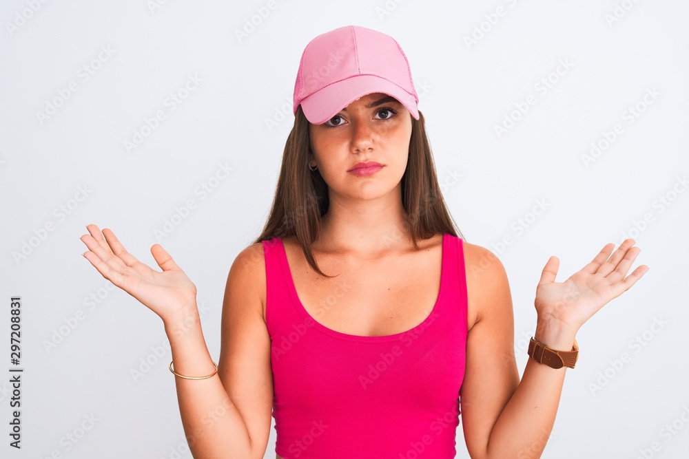 Young beautiful girl wearing pink casual t-shirt and cap over isolated white background clueless and confused expression with arms and hands raised. Doubt concept. Young beautiful girl wearing pink casual t-shirt and cap over isolated white background clueless and confused expression with arms and hands raised. Doubt concept.