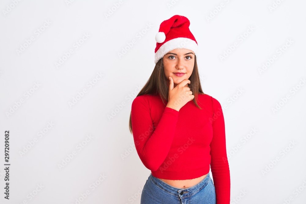 Young beautiful girl wearing Christmas Santa hat standing over isolated white background looking confident at the camera with smile with crossed arms and hand raised on chin. Thinking positive. Young beautiful girl wearing Christmas Santa hat standing over isolated white background looking confident at the camera with smile with crossed arms and hand raised on chin. Thinking positive.