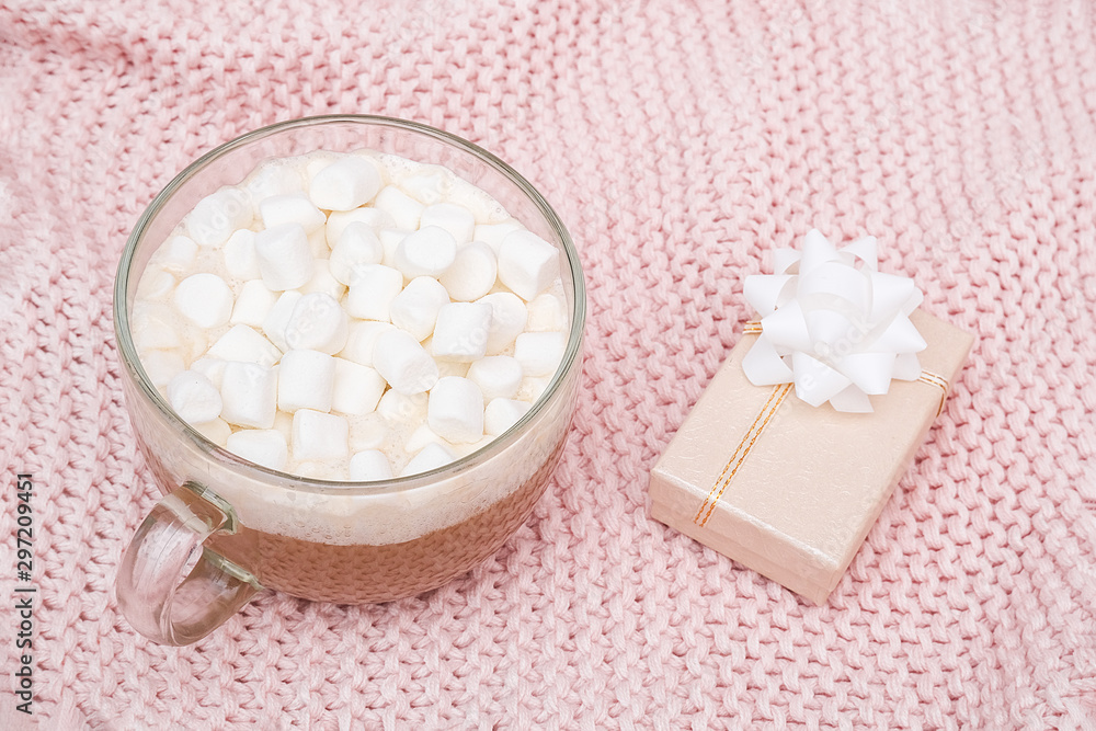 Transparent cup of cocoa with marshmallow and gift box with bow on pink knitted plaid, close up Transparent cup of cocoa with marshmallow and gift box with bow on pink knitted plaid, close up