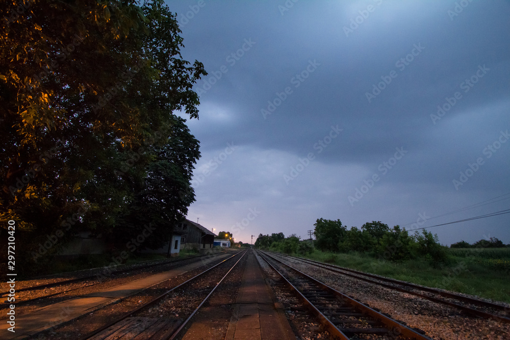 Fototapeta premium Railway tracks, rails and platforms in a rural train station in Alibunar, Serbia, taken during sunset, after a cloudy and rainy storm with trees and a perspective