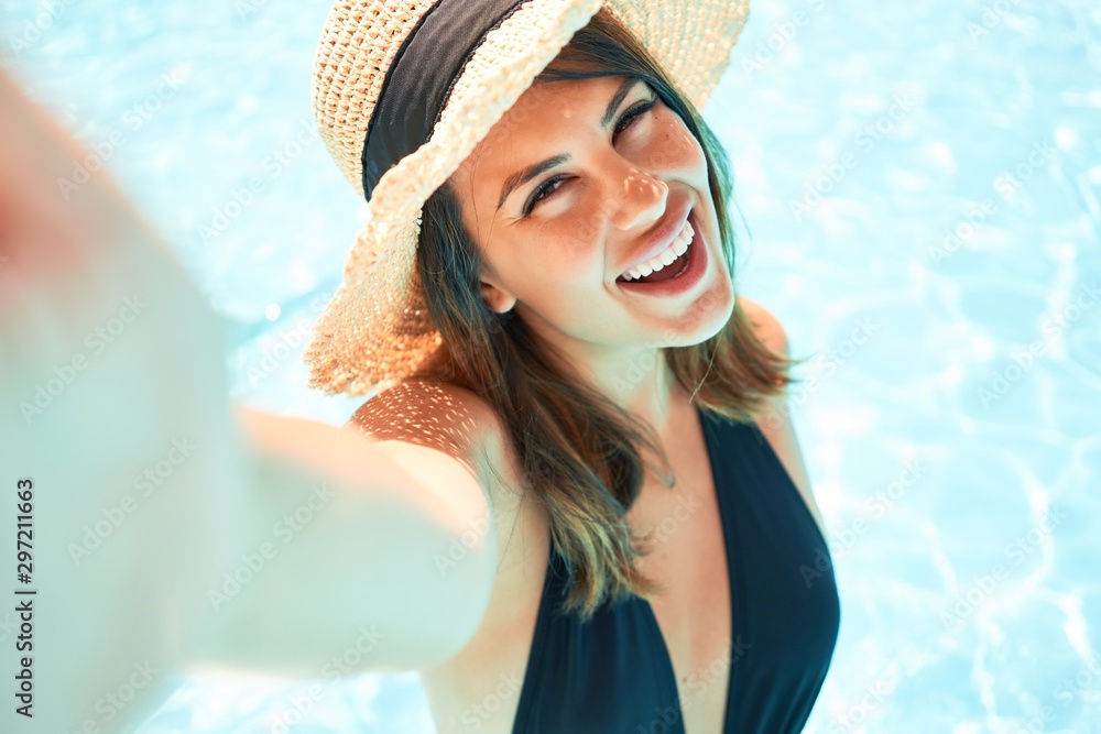Young beautiful and sexy woman at the hotel pool taking a selfie smiling using smartphone on sunny day - obrazy, fototapety, plakaty 