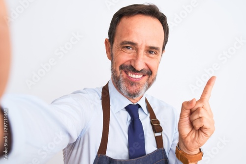 Middle age shopkeeper man wearing apron make selfie over isolated white background very happy pointing with hand and finger to the side