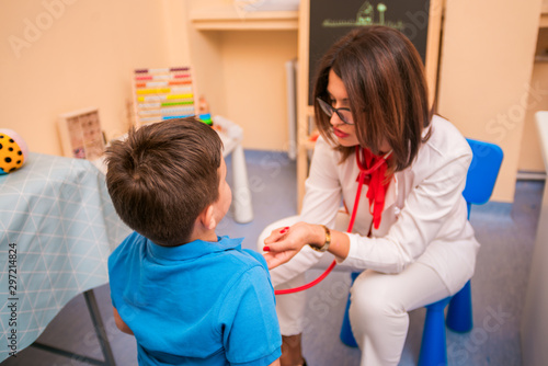 Portrait of adorable little boy visiting doctor  looking brave and smiling  holding while pediatrician listening to heartbeat with stethoscope